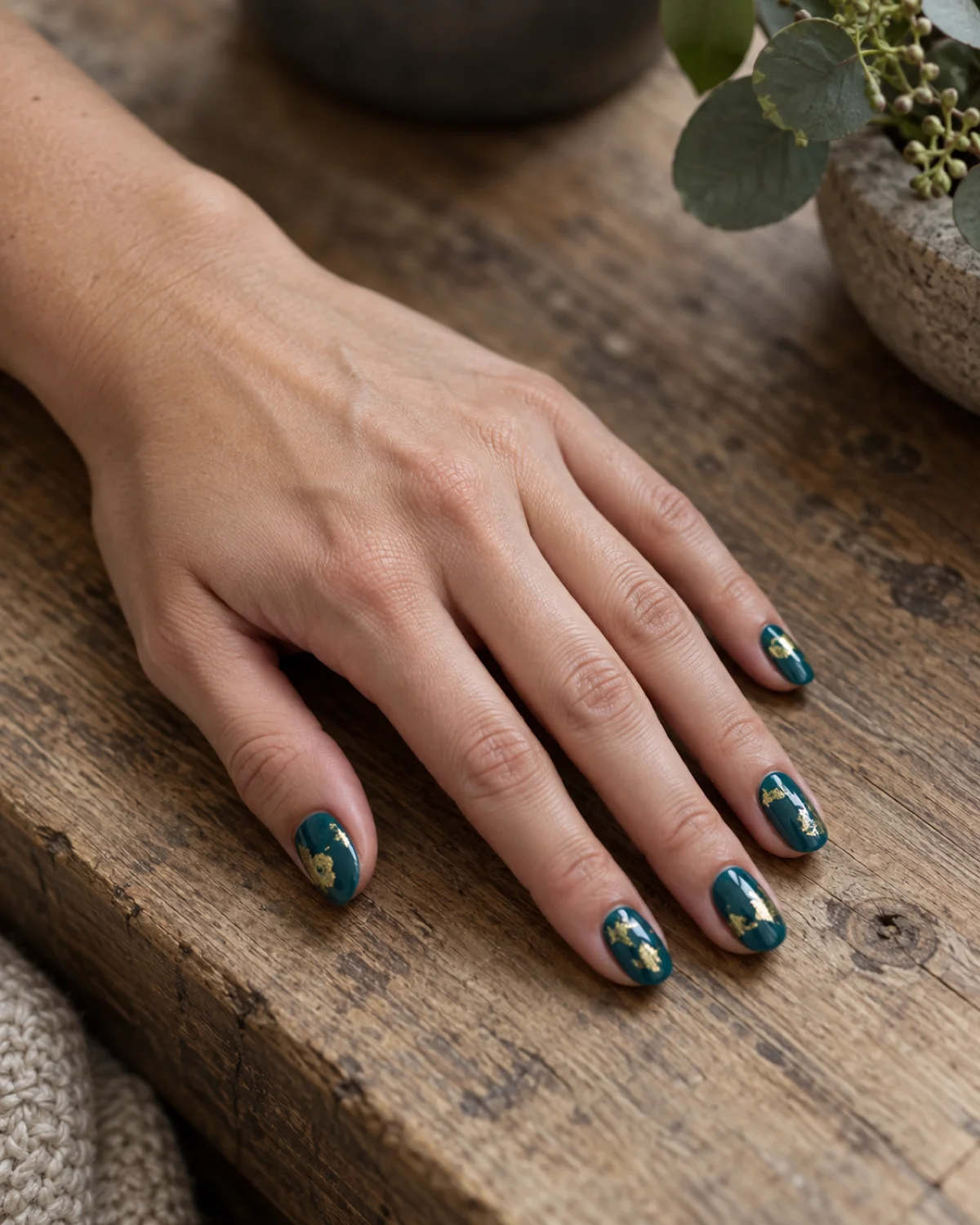 Single left hand with oval nails in rich teal gel with gold leaf flecks resting palm down on raw wood surface