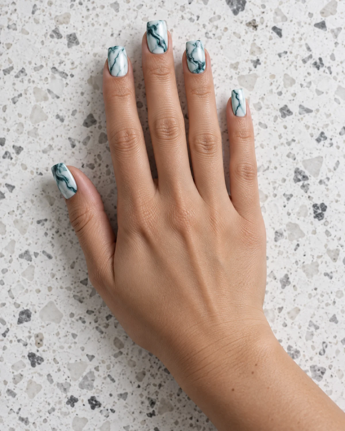 Single right hand with square nails showing teal and deep teal marble veining on white base resting palm down on white terrazzo surface