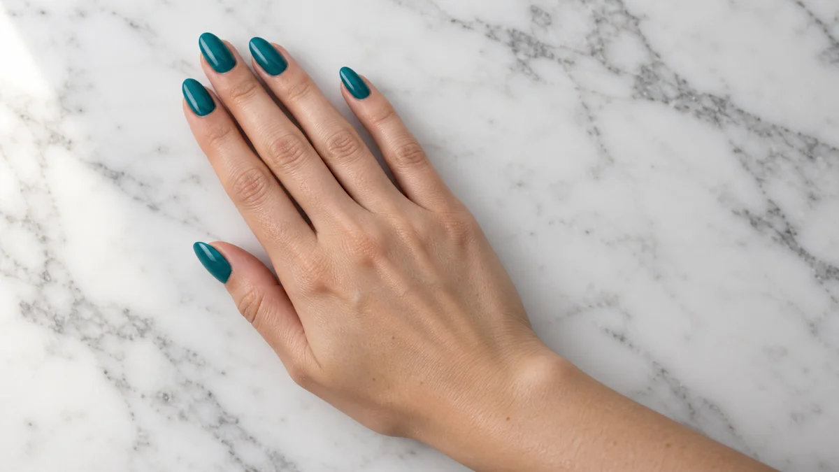 Single right hand with almond nails in rich mid-teal crème polish resting palm down on white marble countertop