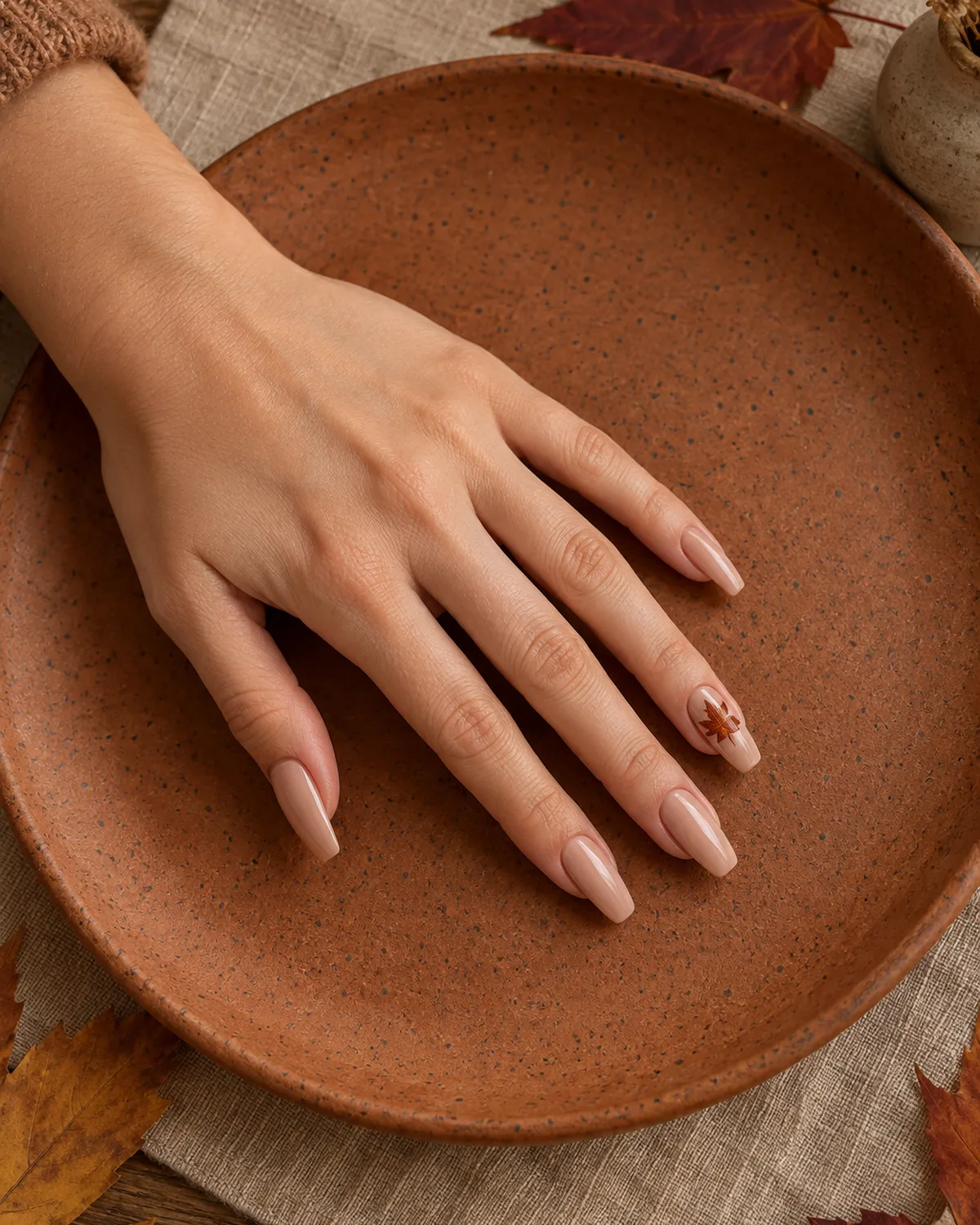 Warm nude coffin nails with tiny maple leaf accent painted on ring finger in burnt sienna and gold