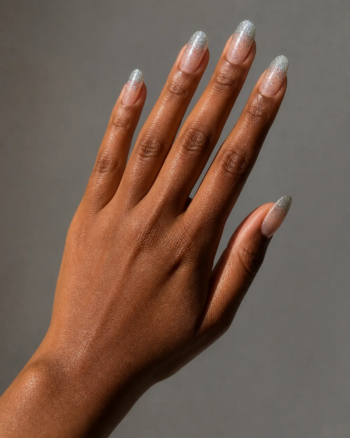 Single left hand reaching upward on a diagonal with medium almond nails featuring glitter fade from sheer to dense silver sparkle