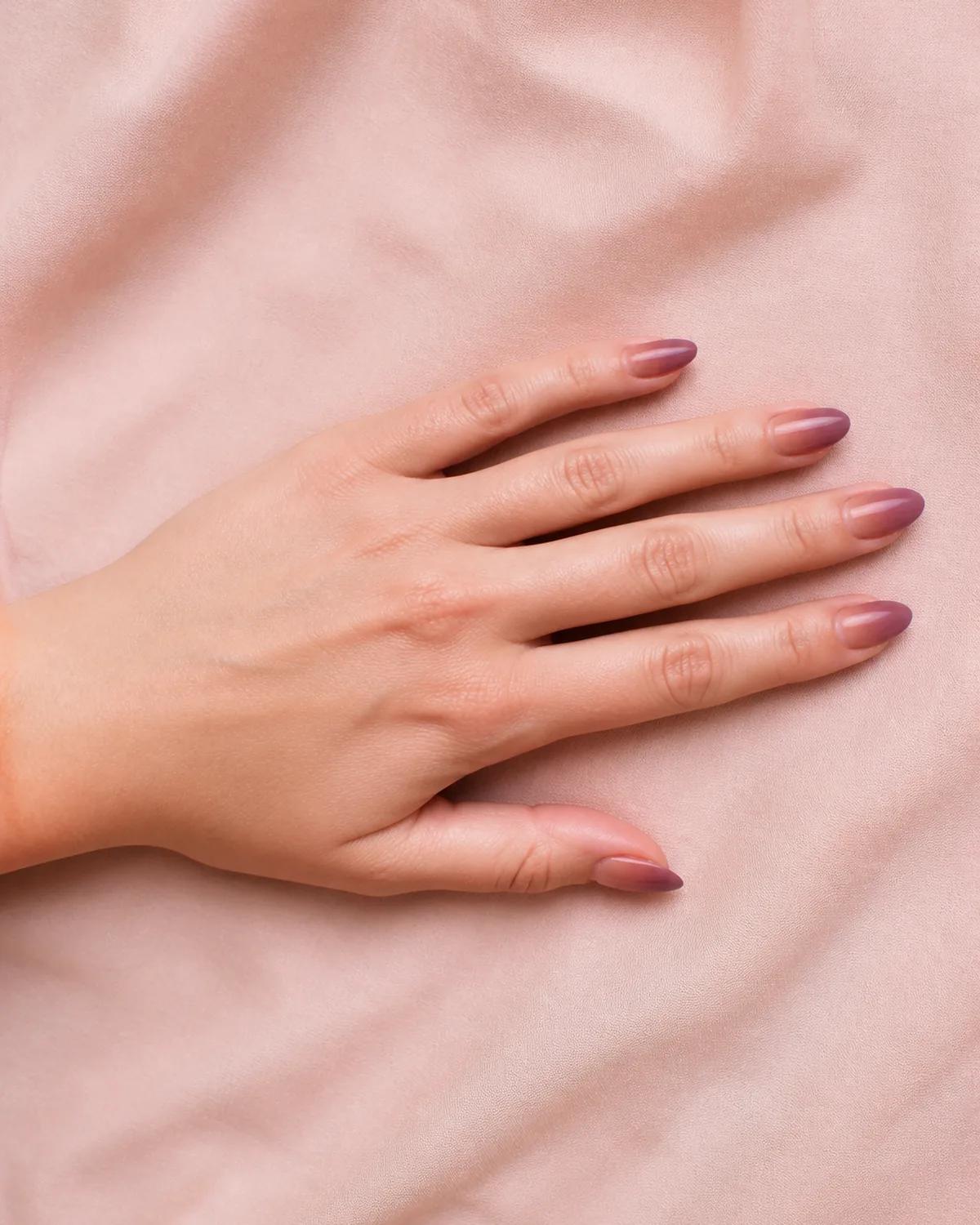 Single left hand lying flat on blush fabric with medium almond nails featuring dusty rose-to-mauve two-tone gradient