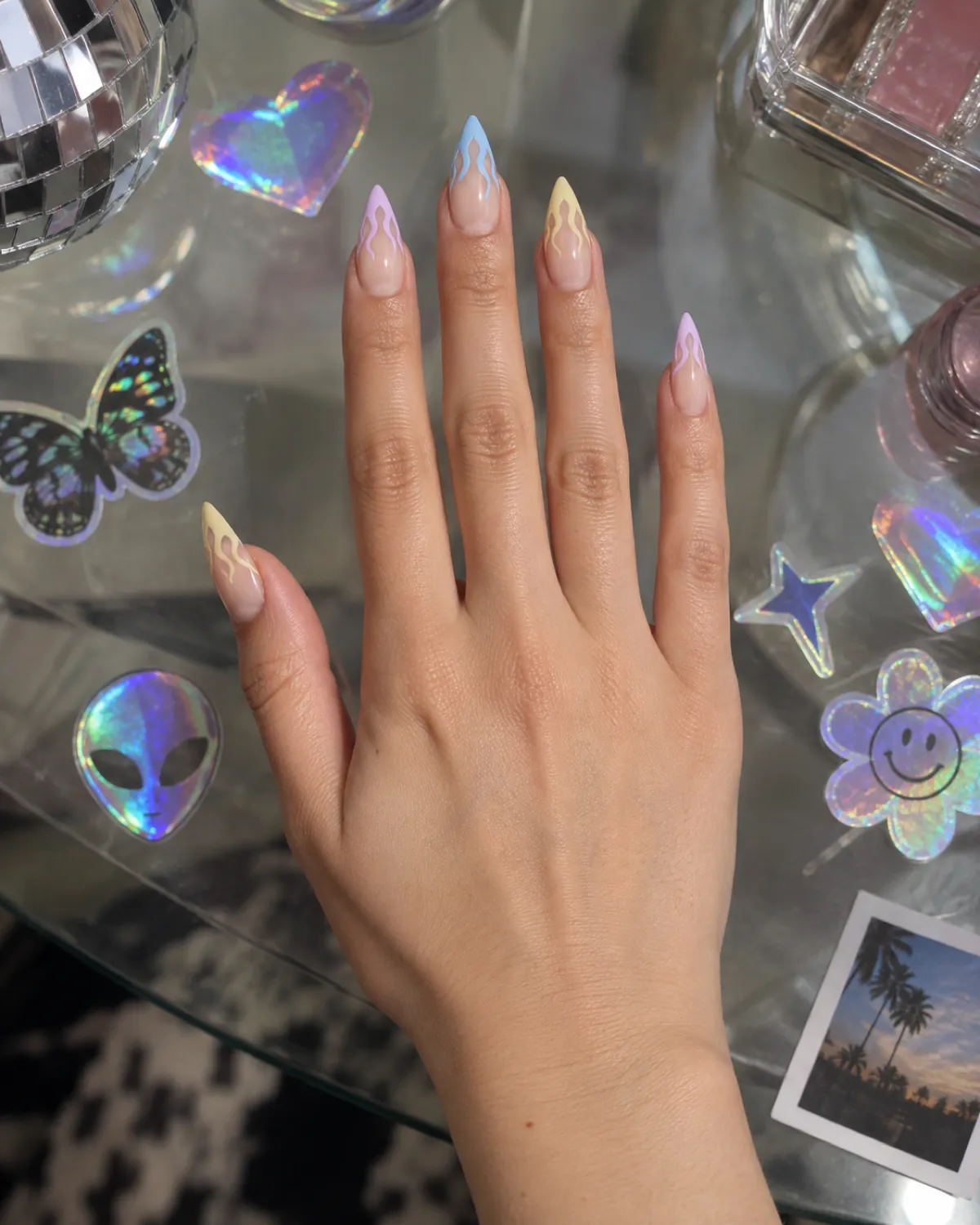 Single right hand with stiletto nails featuring pastel flame tips palm-down on clear glass tabletop with holographic surface