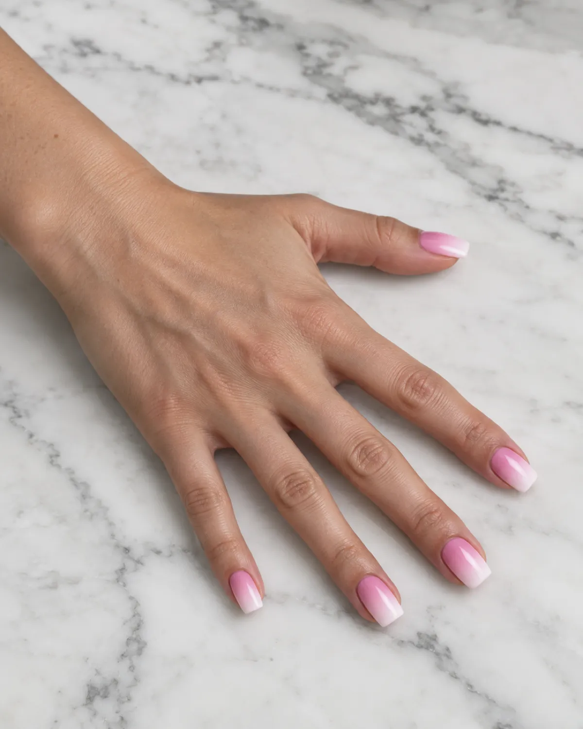 Single left hand with square nails in airbrushed candy pink to white gradient palm-down on white marble countertop