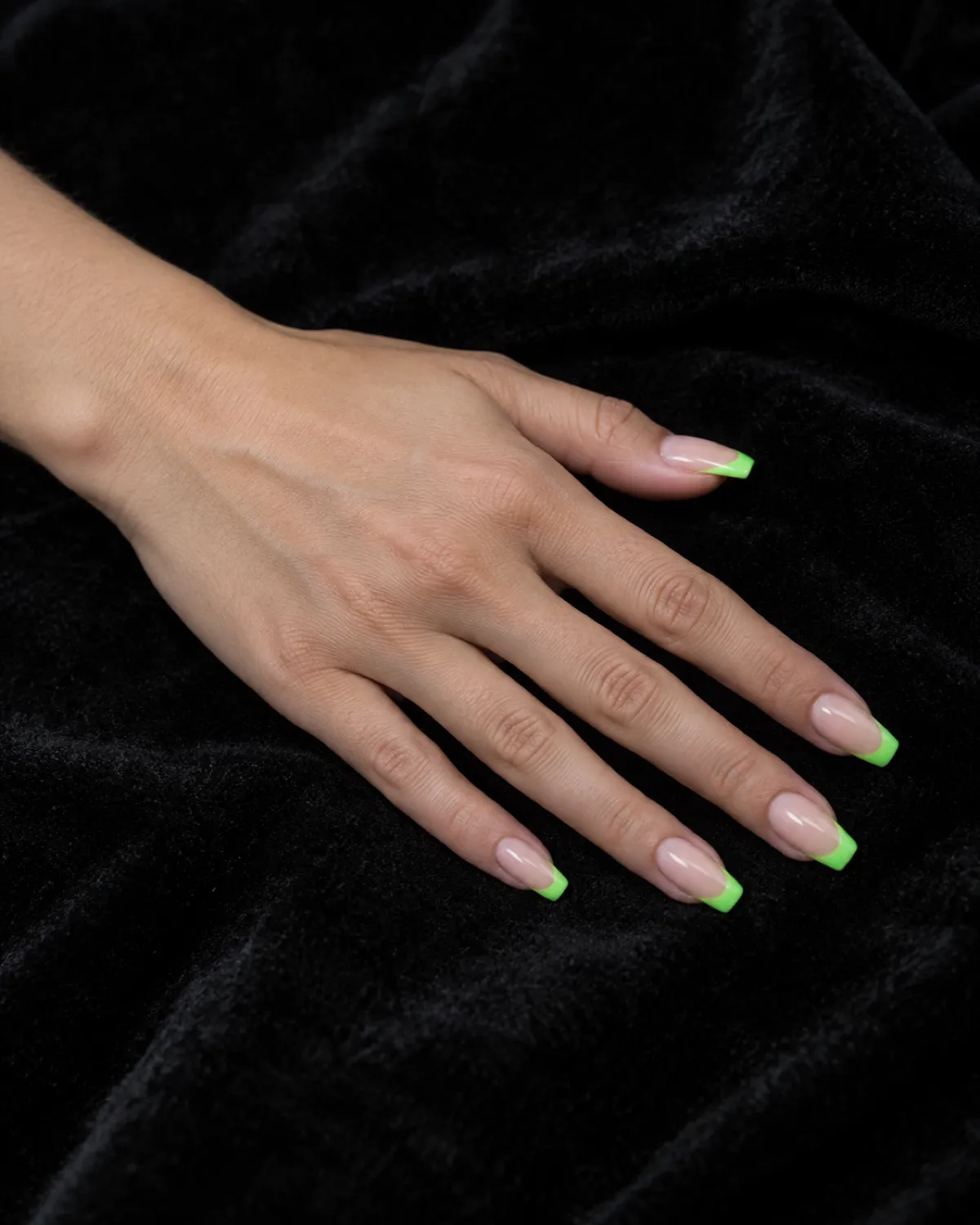 Single left hand with coffin nails featuring neon green French tips palm-down on black velvet surface